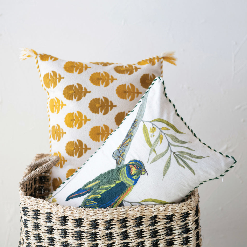 white pillow with blue and green embroidered bird standing on a branch in basket with other decorative pillow.