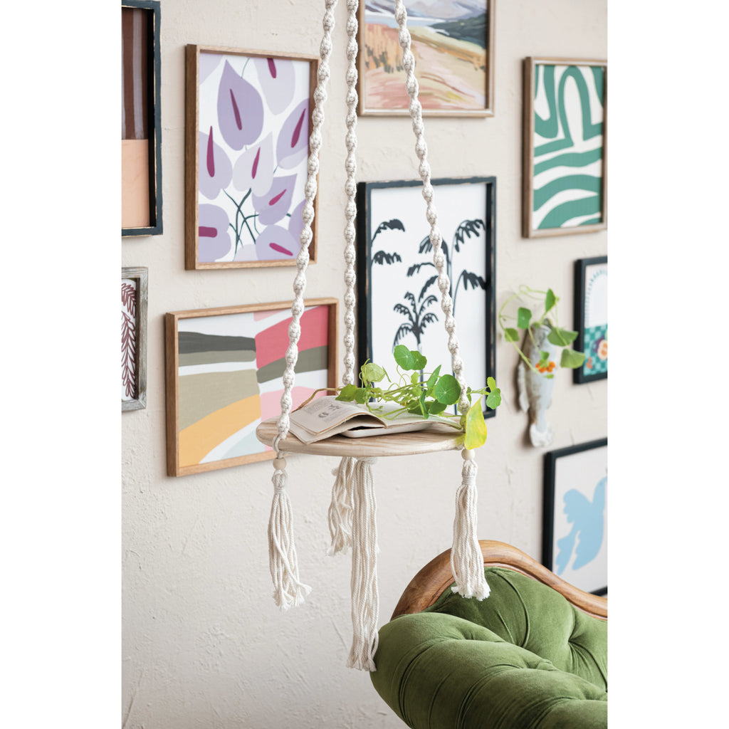 Wood & Cotton Macrame Plant Hanger/Shelf with book and plant cuttings  in front of wall with abstract floral prints and green couch