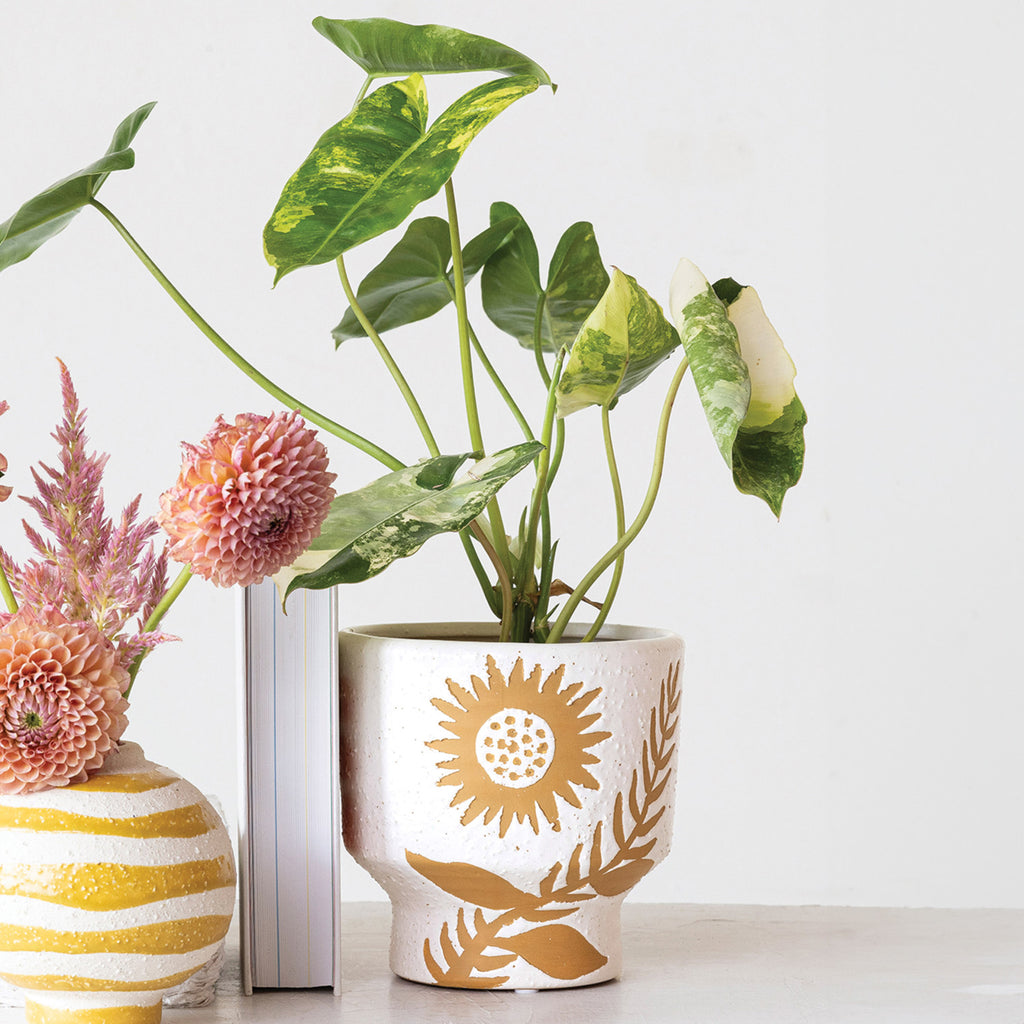 White glazed footed planter with yellow undercolor showing flower and leaves with plant inside, and vase with dahlias in background