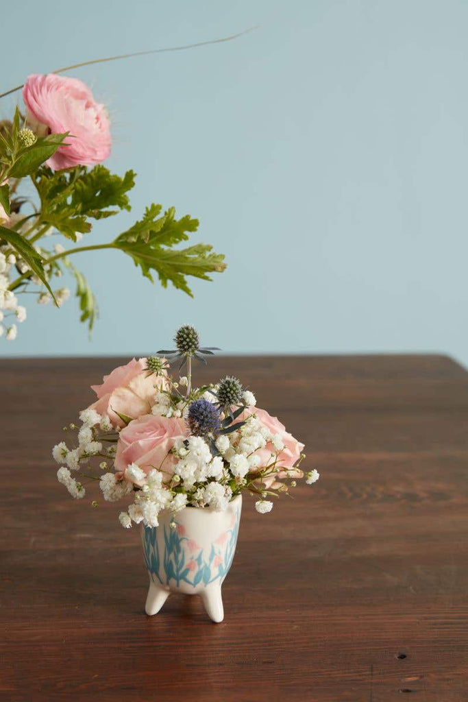 Floral Spring Footed Pot with blue leaves and pink flowers painted on. In front of blue backdrop on wooden table.